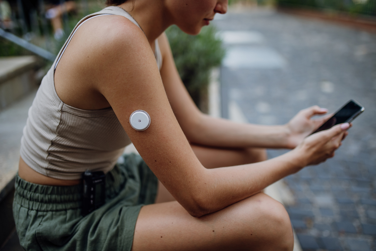 Une personne est assise dehors. Elle a un capteur rond sur le haut du bras. Elle regarde son téléphone portable. Scanner le capteur avec son téléphone portable lui permet de mesurer la quantité de sucre dans son sang.